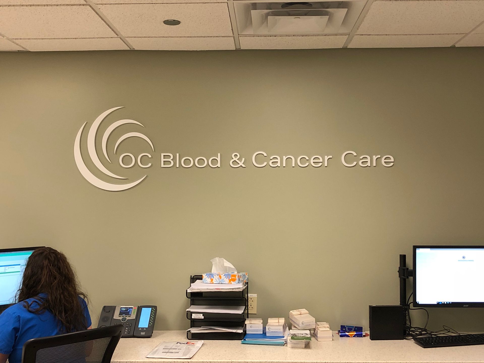 A woman is sitting at a desk in front of a sign that says oc blood and cancer care.