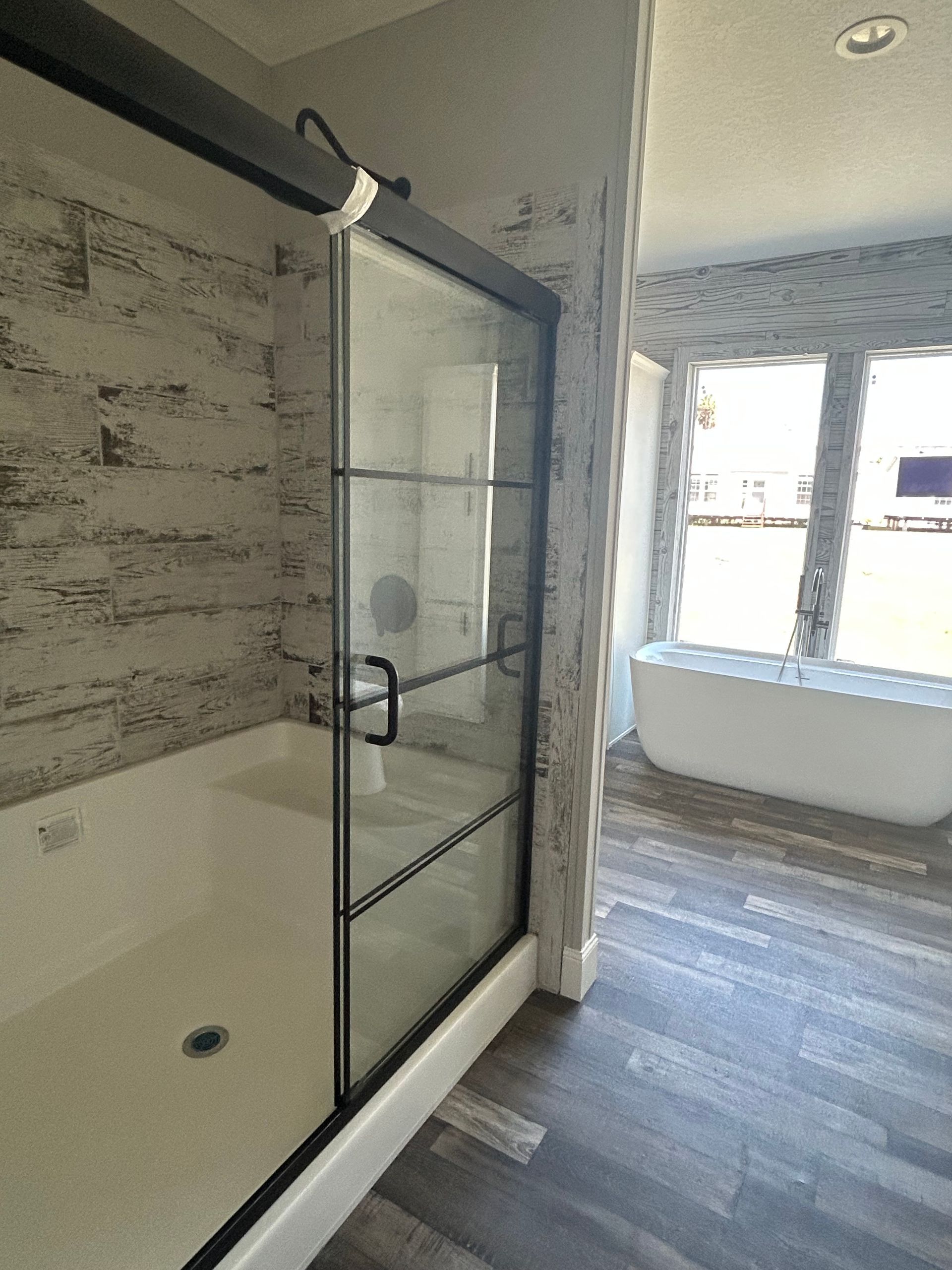 A modern bathroom with a glass shower, white bathtub, and dark wood-look floors. Textured white wall and natural light from windows.
