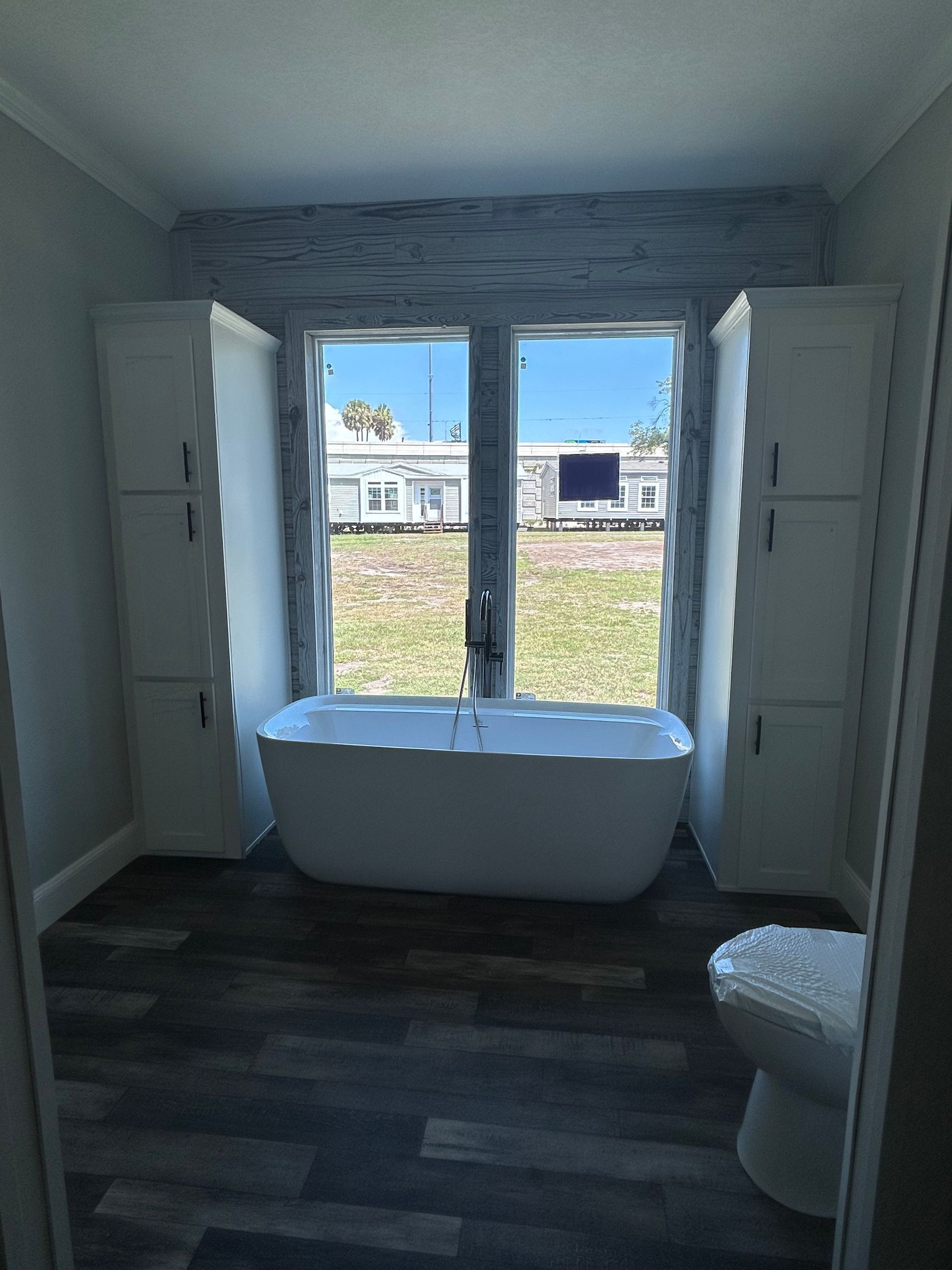Bathroom with a freestanding white tub in front of a window, flanked by tall white cabinets on a dark wood-look floor.