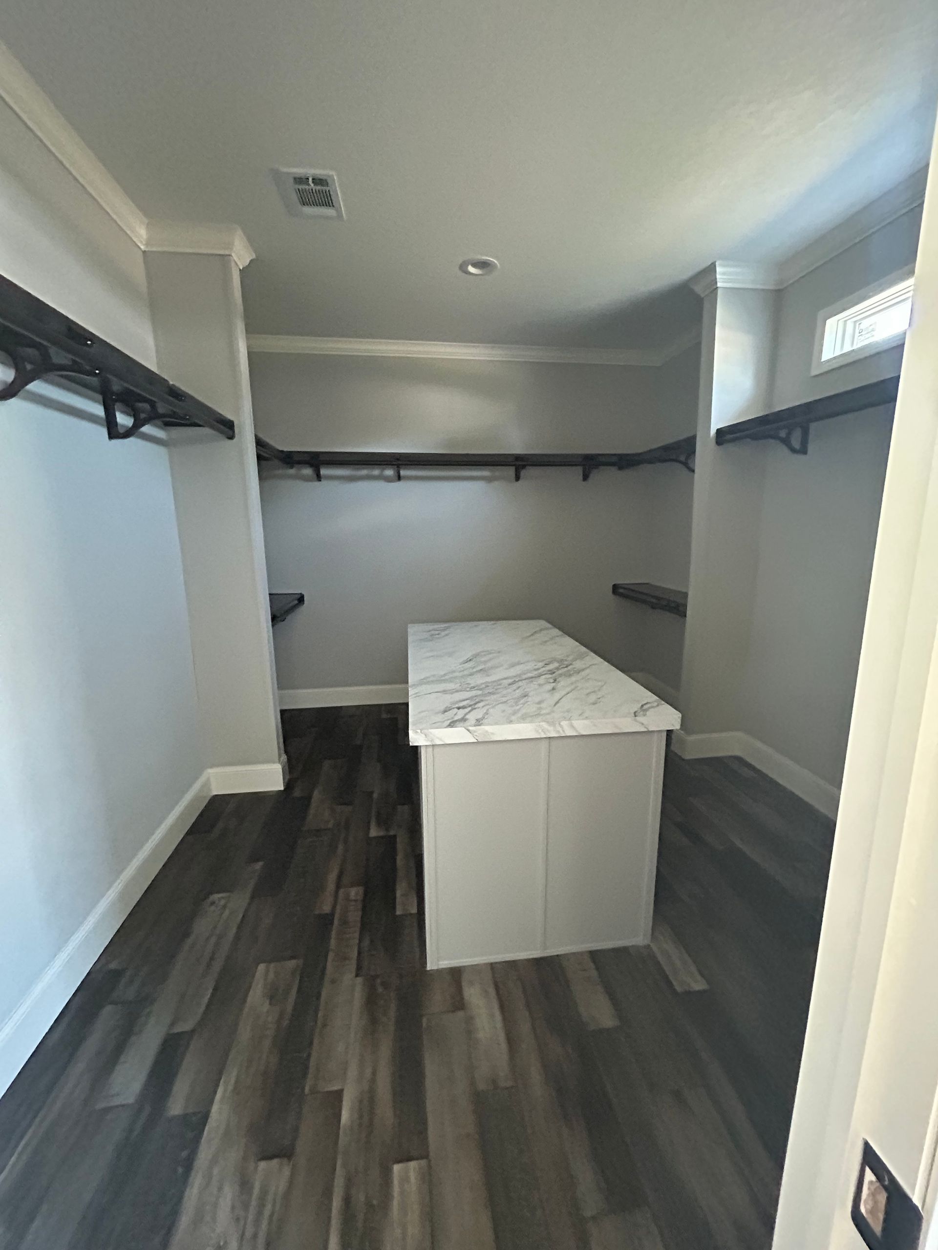 A walk-in closet with gray walls, dark wood-look flooring, and a center island with a white countertop. Clothing rods line the walls.