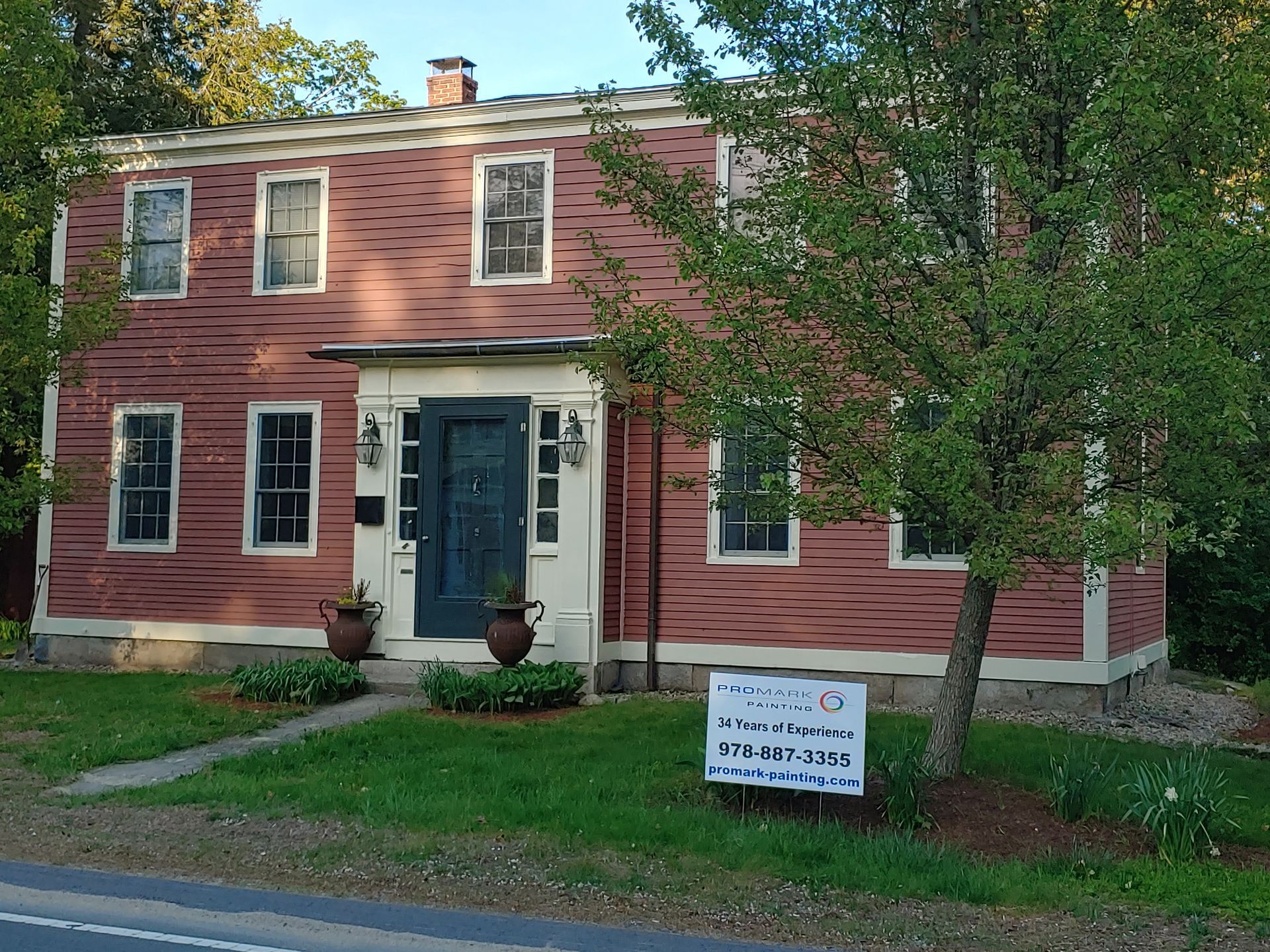 A two-story, reddish-brown clapboard house with white trim, a dark front door, and a sign in the front yard.
