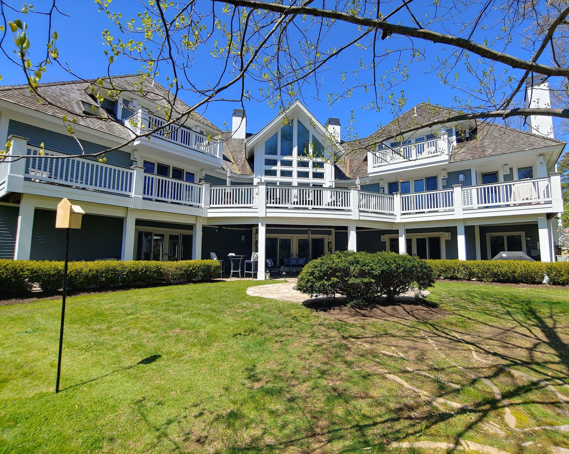 A large, multi-story blue house with white trim, balconies, and a landscaped lawn on a sunny day.