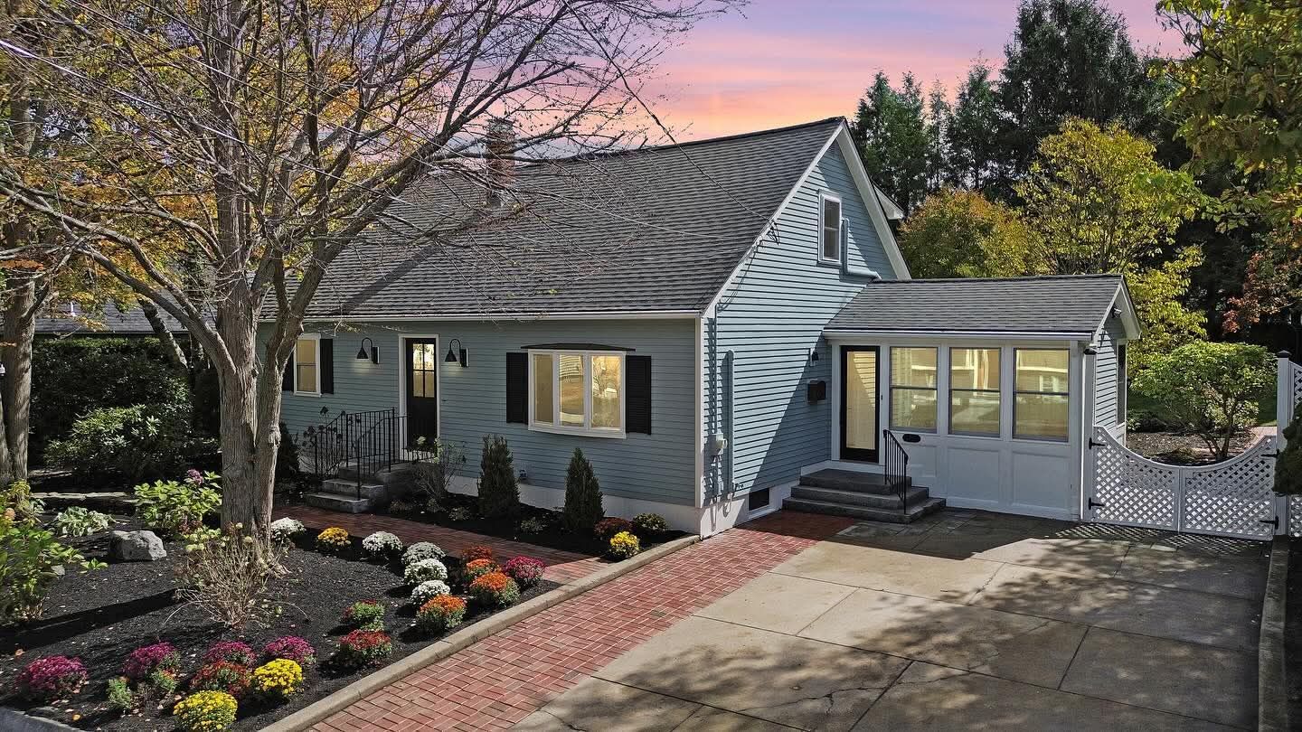 Blue house with black trim and brick driveway, surrounded by fall foliage.