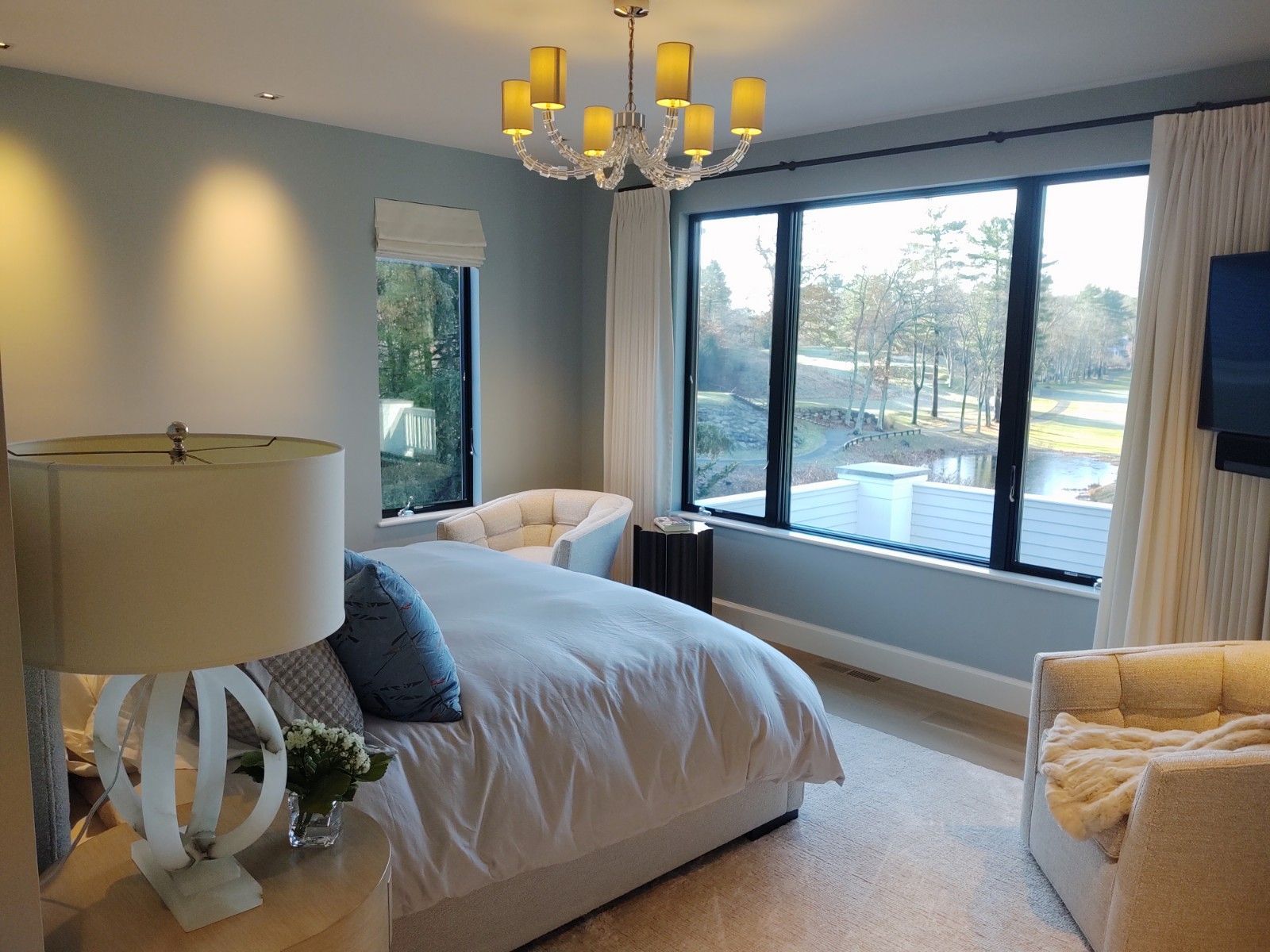 Bedroom with bed, windows, armchairs, and a chandelier; light blue walls, neutral carpet, outdoor view.