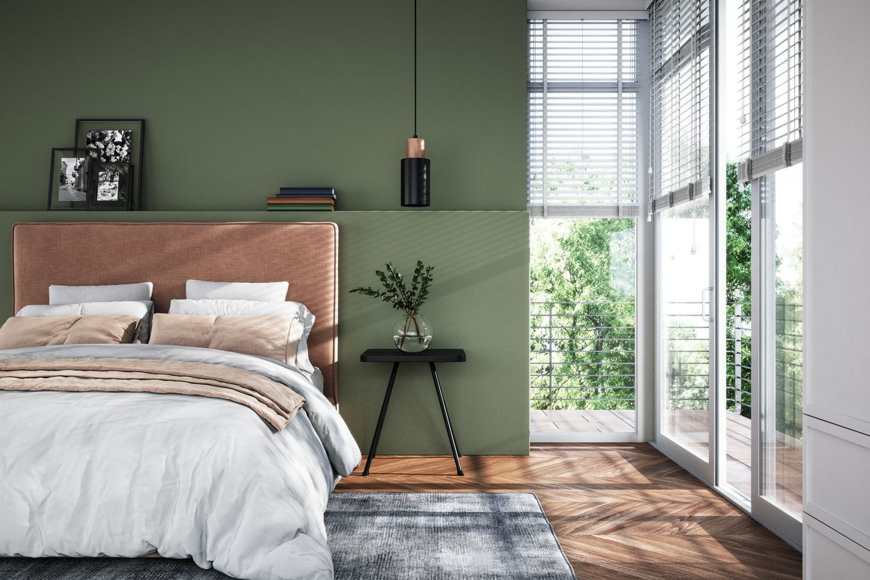 Bedroom with olive green wall, bed with tan headboard, large windows, and sunlight.