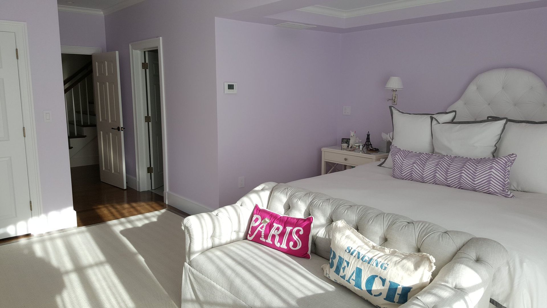 Bedroom with lilac walls, tufted headboard, and bench with pillows. Sunlight streams in.