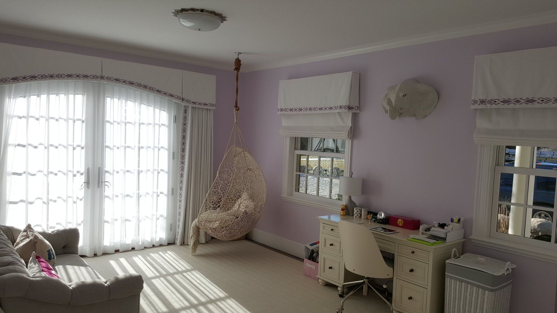 Girl's bedroom with lavender walls, white trim, a hanging chair, desk, and windows with sheer curtains.