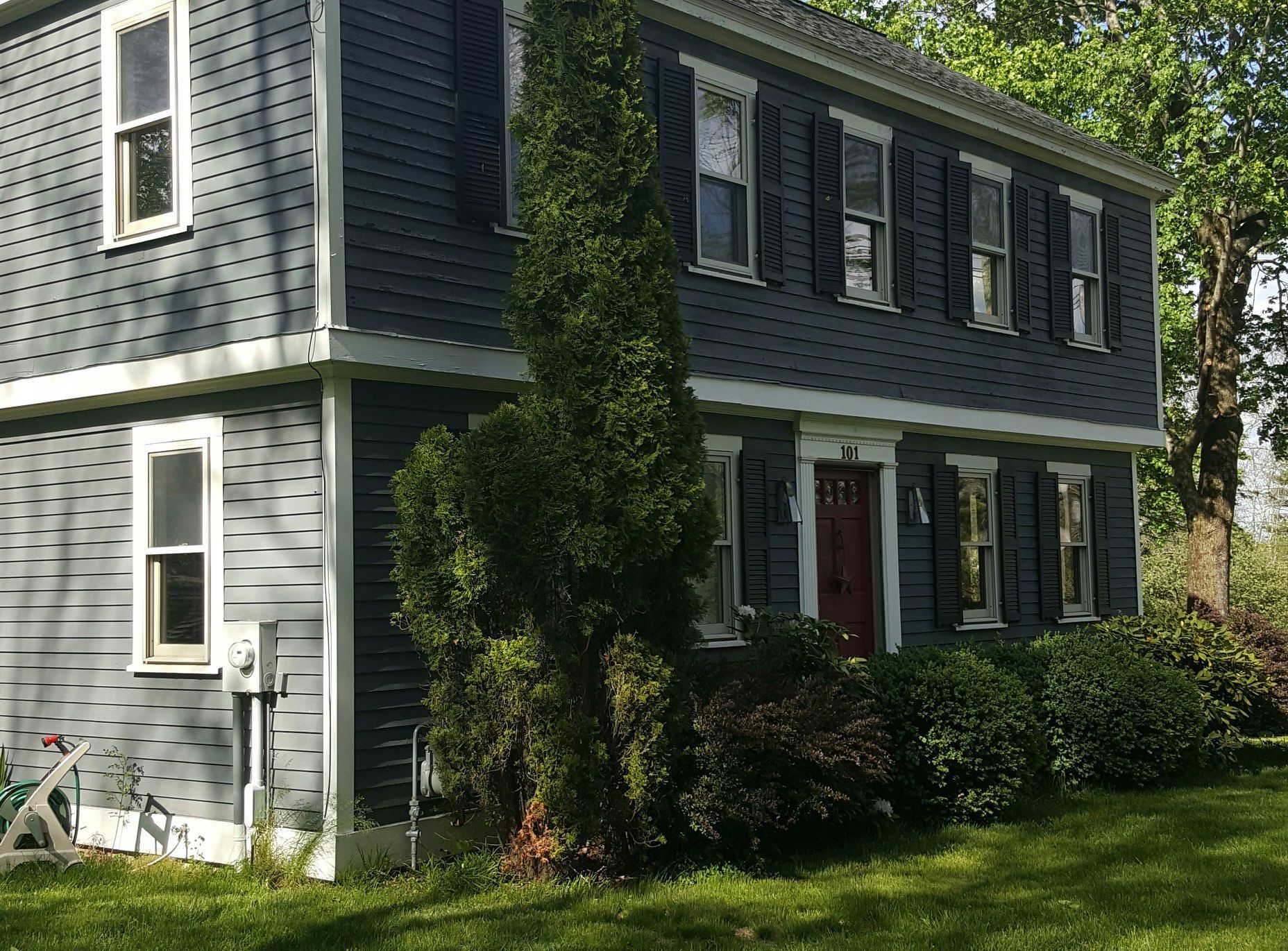 Two-story blue house with white trim, black shutters, and a red door. A large evergreen tree is in the front yard.