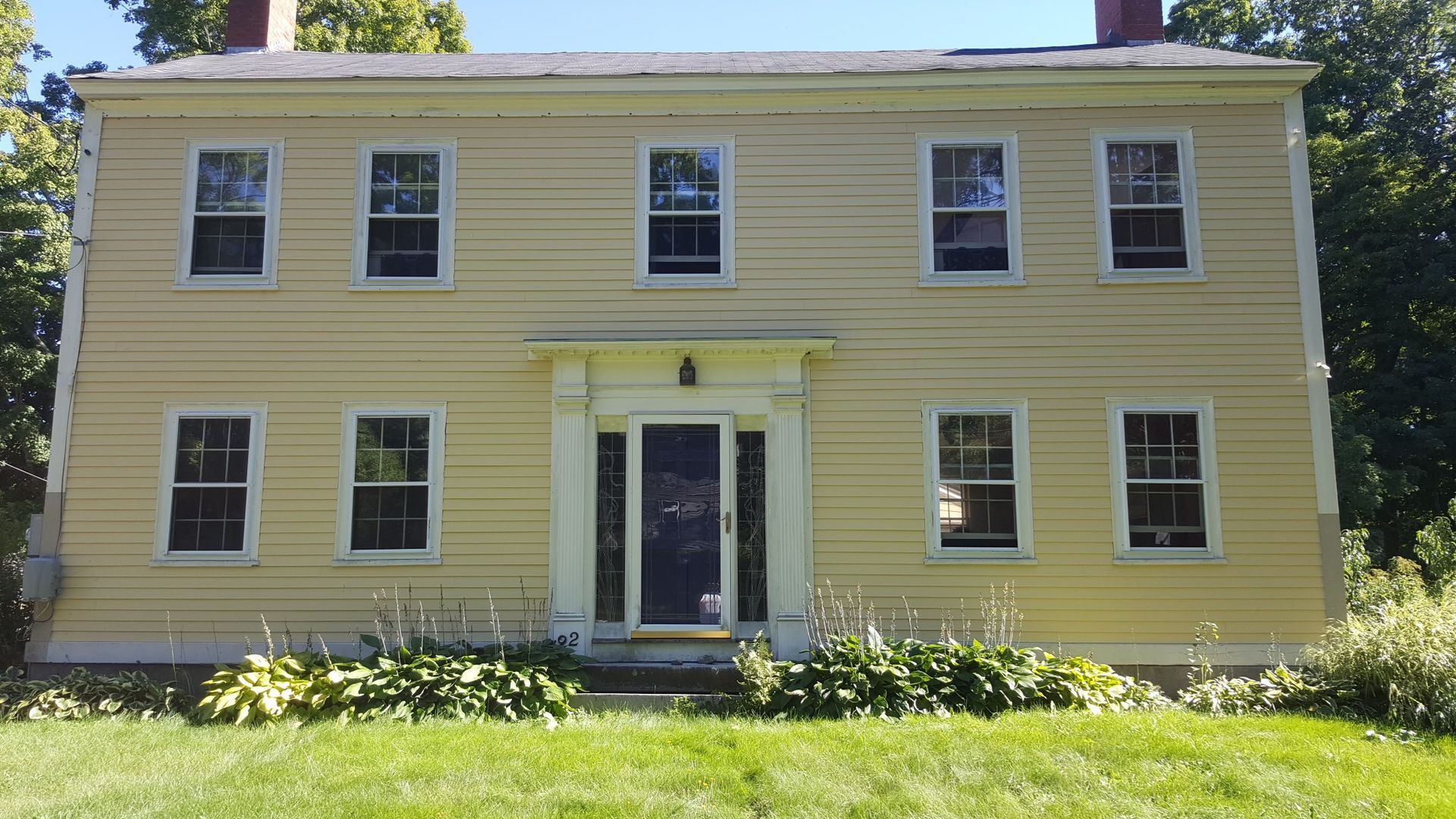 Yellow two-story house with black-screened door, eight windows, and two red chimneys. Green grass and foliage in front.