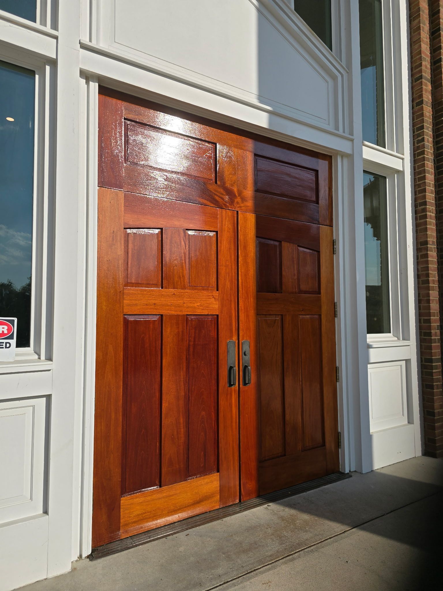 Two glossy brown wooden doors with rectangular panels, set in a white frame, flanked by windows.