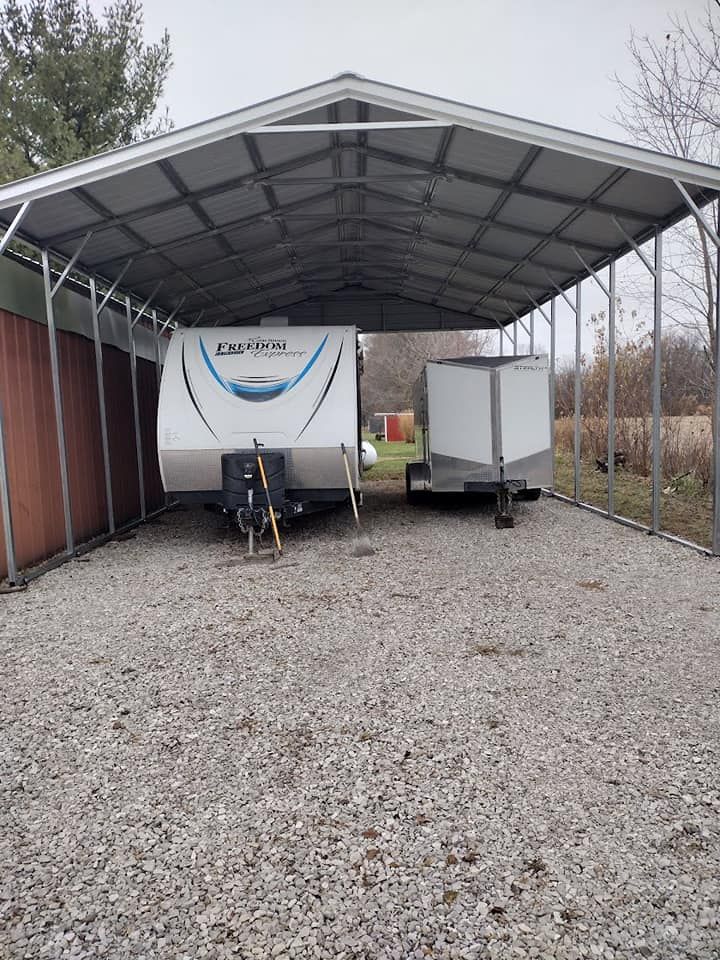 A camper and enclosed trailer parked under a metal carport on gravel.