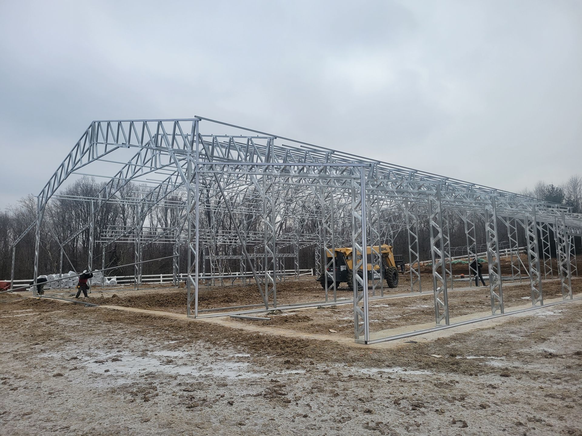 Metal frame of a building under construction on a snowy field.  A tractor and workers are visible.