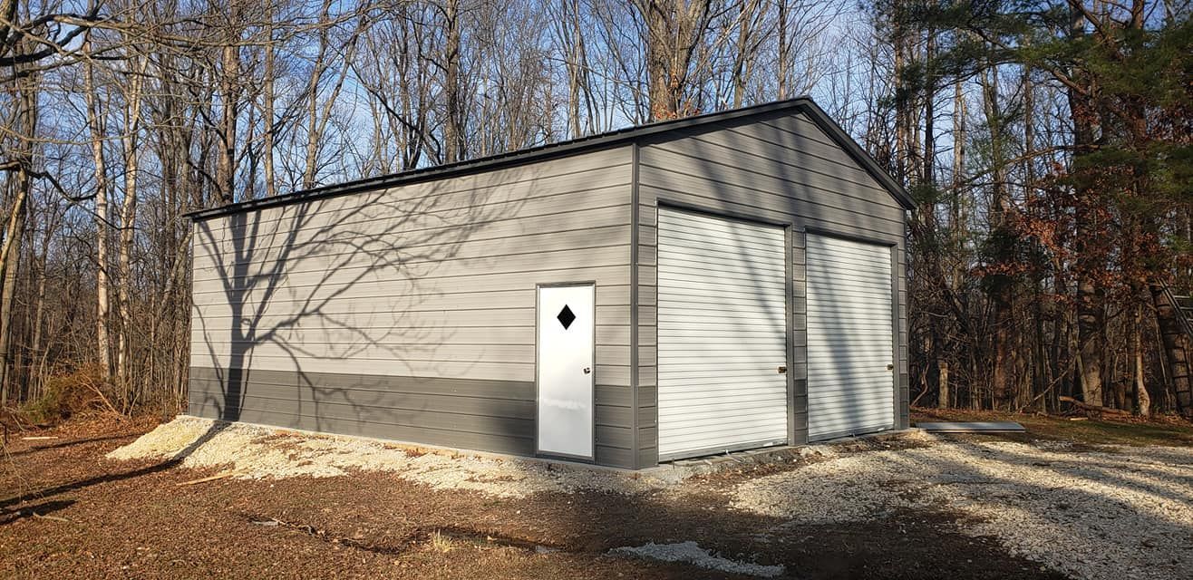 A gray metal garage with two doors and a side door is set in a wooded area with gravel.