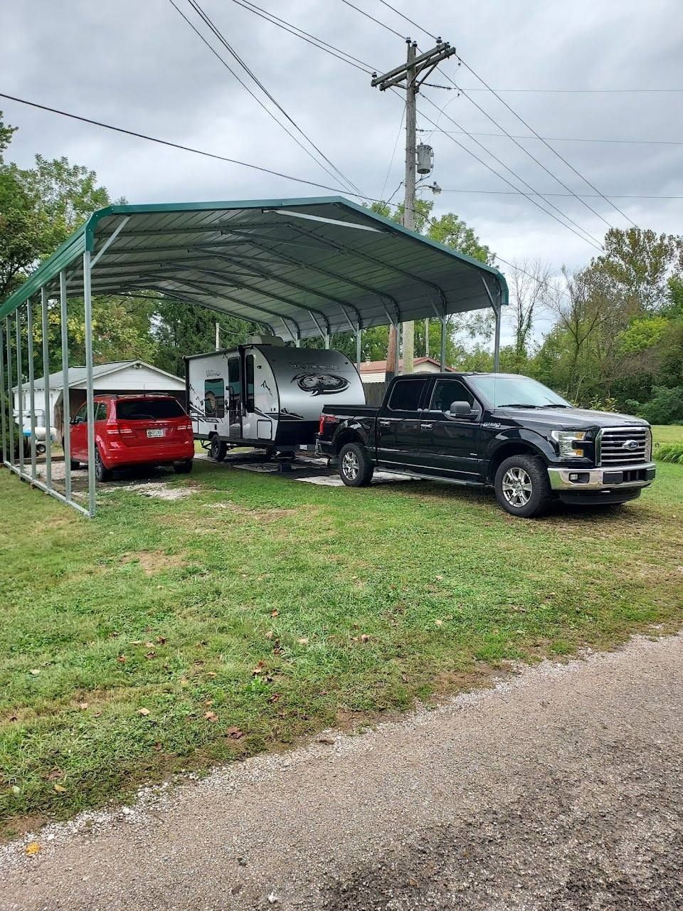 Black pickup truck towing a camper under a green carport, red car parked beside.