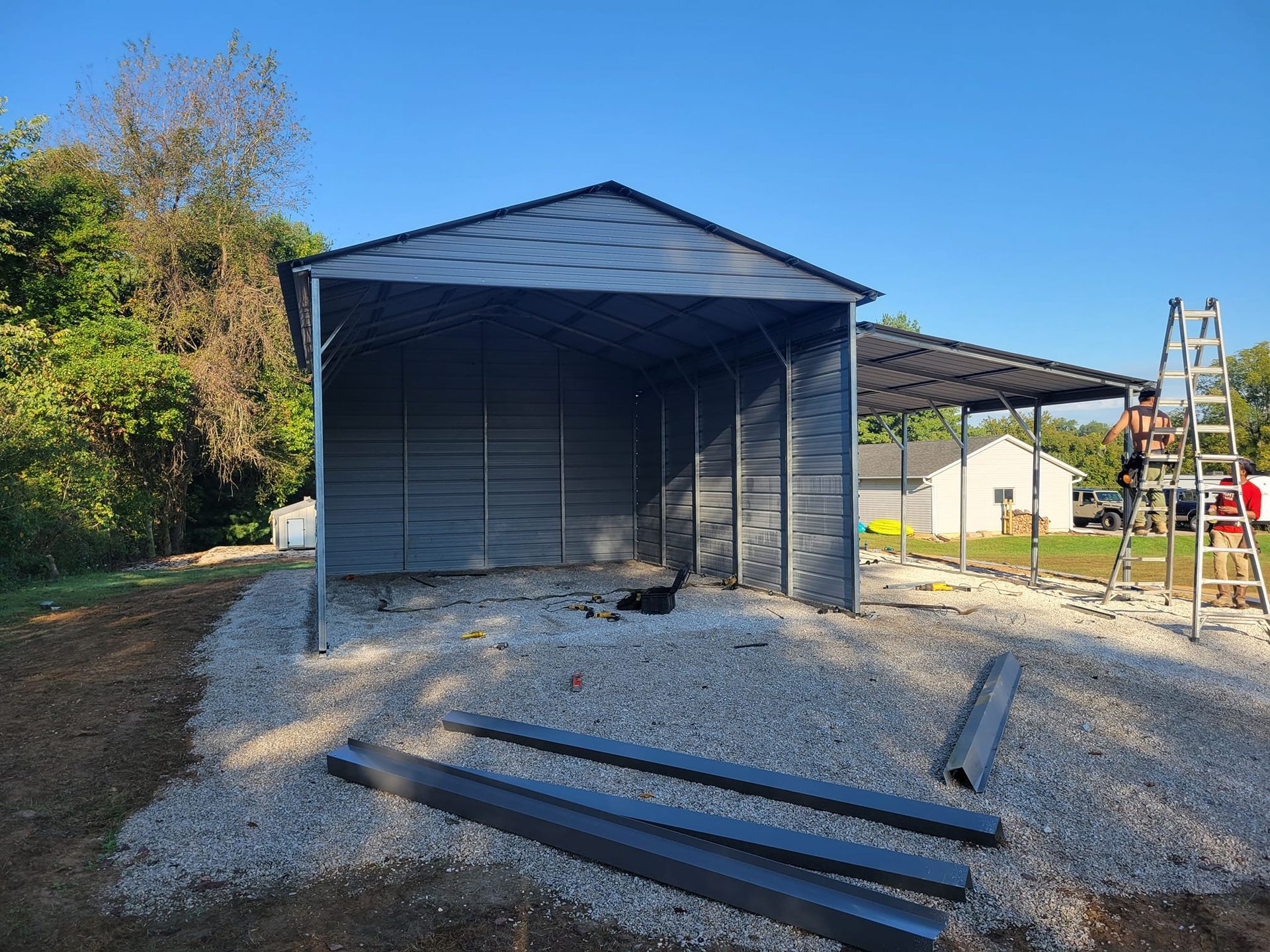A partially-constructed gray metal shed on gravel; a person on a ladder works nearby.