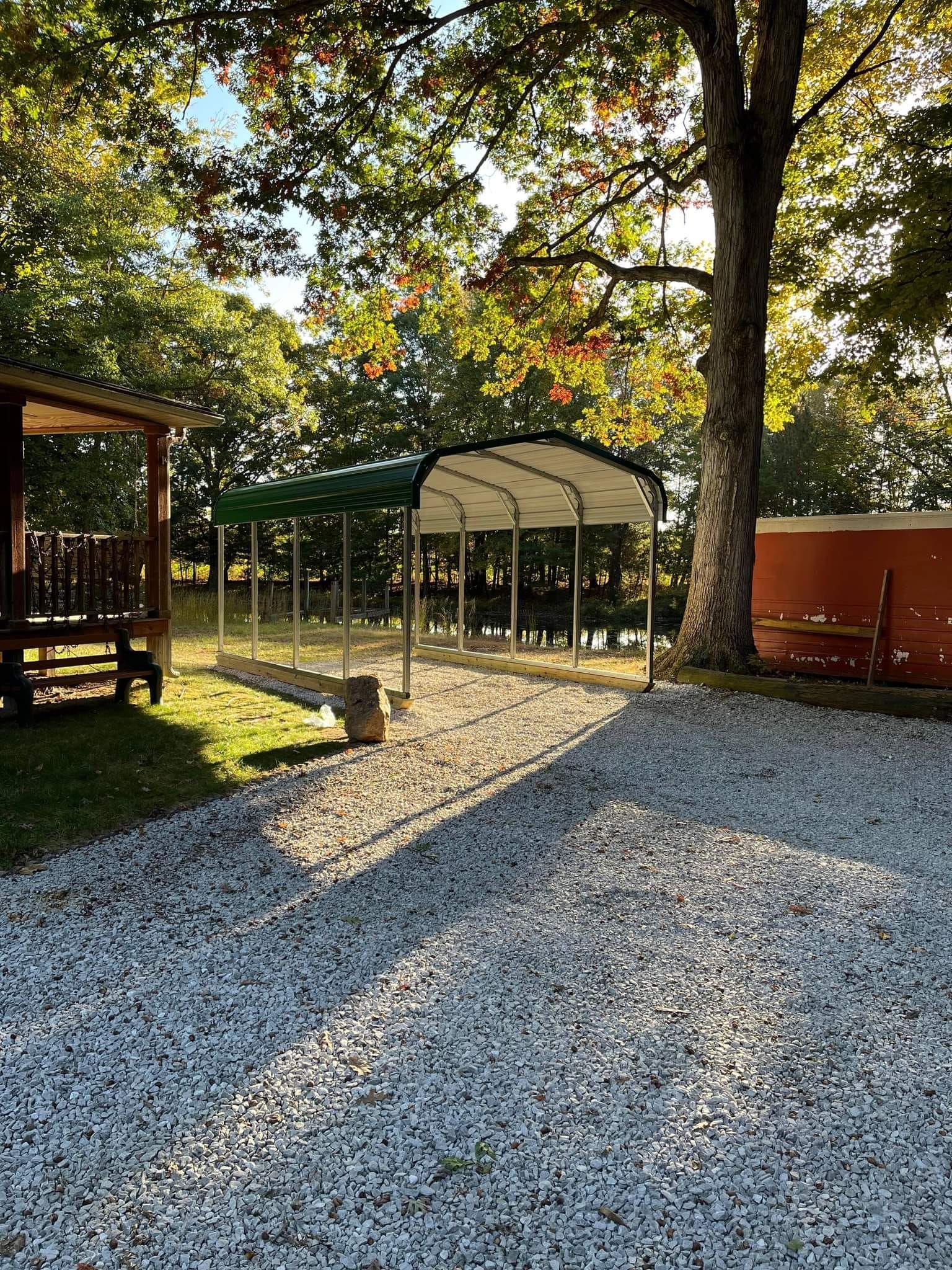 Gravel driveway leads to a fenced enclosure under a green-roofed shelter, with a porch and hot tub visible.