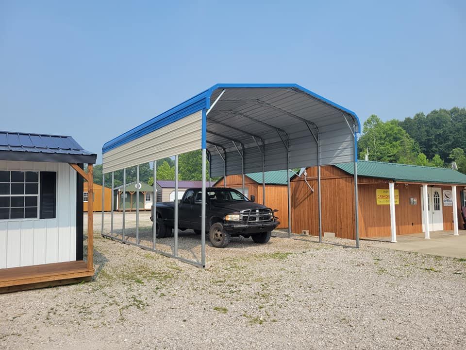 Black pickup truck parked under a blue and beige metal carport in a gravel lot.