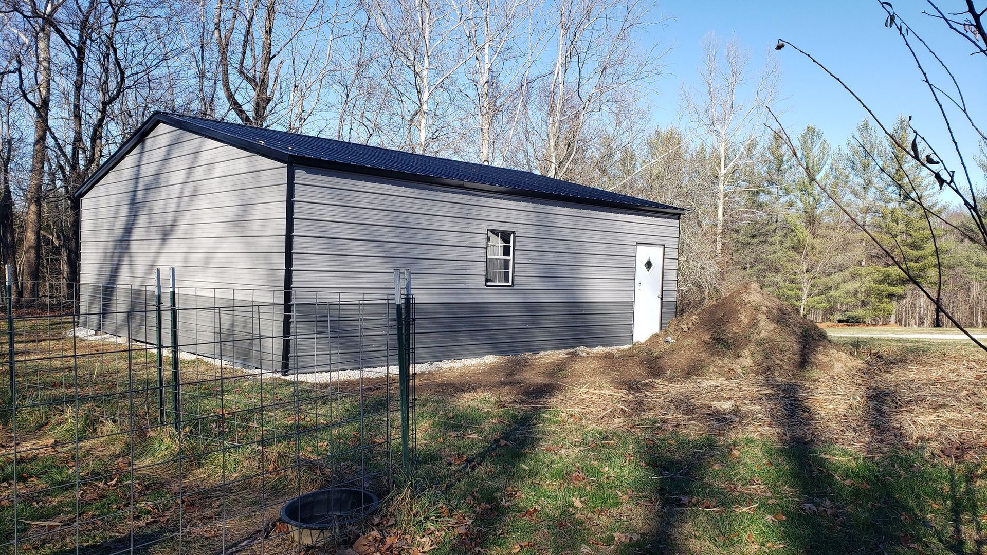 Gray shed with black roof in a wooded area. Fenced area in front.
