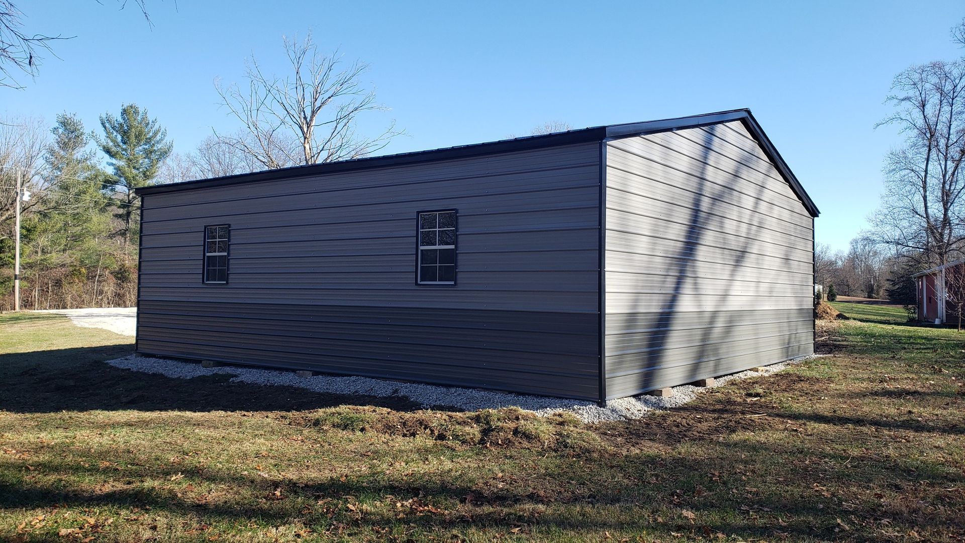 Gray metal building with dark roof and two windows on a grassy lot under a blue sky.