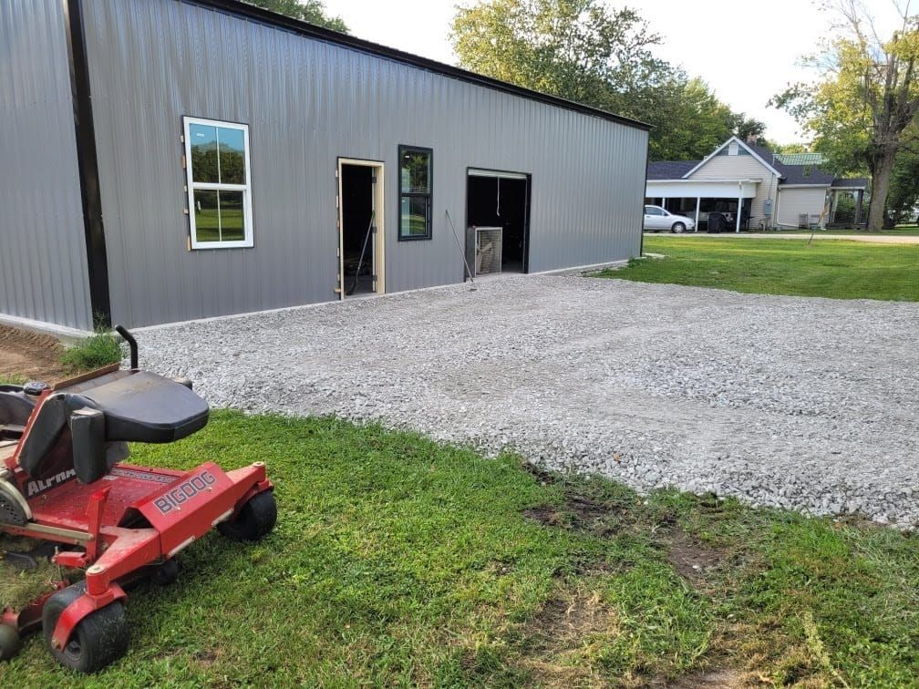 Gray metal building with gravel driveway and lawn mower on grass. A house is visible in the background.