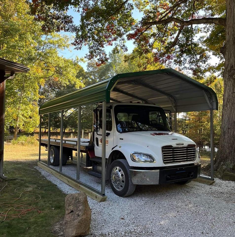 White truck parked under a green metal carport on a gravel driveway, in a wooded area.