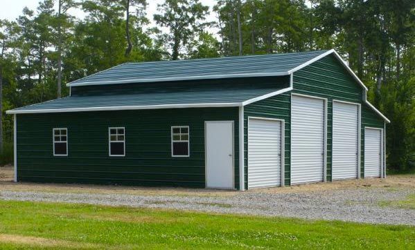 Green metal barn with white doors and windows; gravel driveway, grassy field, trees in background.