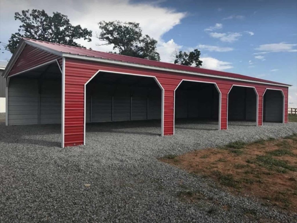Red and gray metal carport with four open bays on gravel, under a blue sky.
