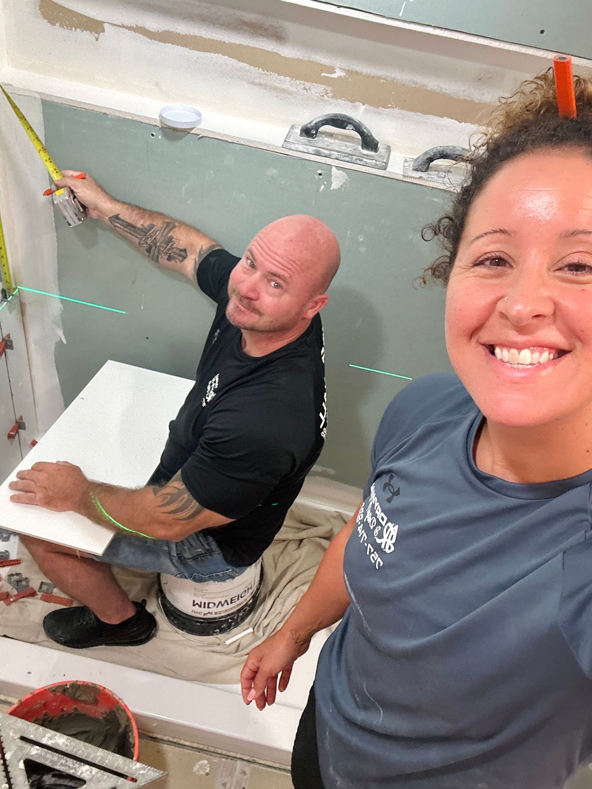 Two people working on tiling in a bathroom. Man measures; woman smiles, taking a selfie.