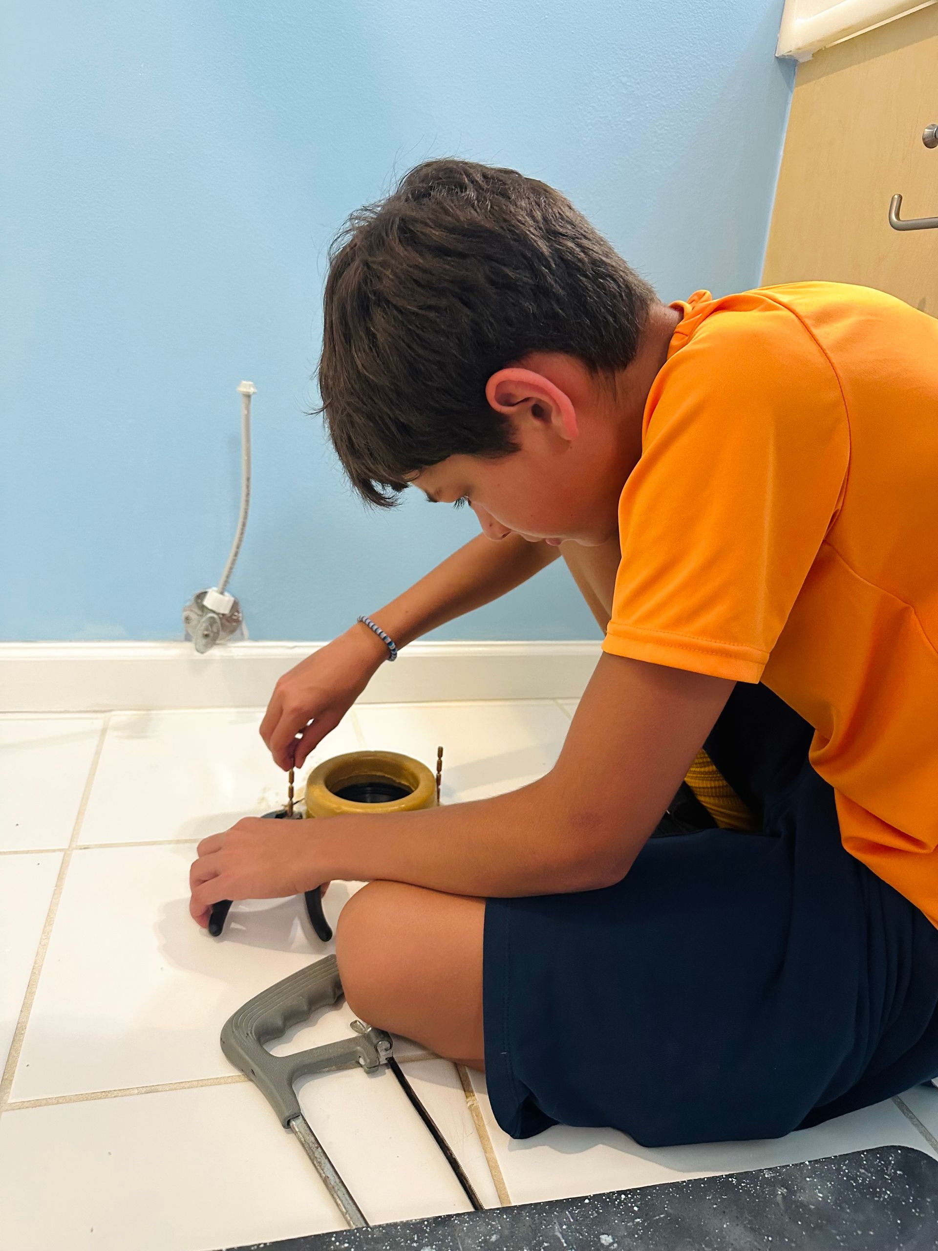 Boy in orange shirt kneels, working on a toilet. Blue wall, white tile floor.