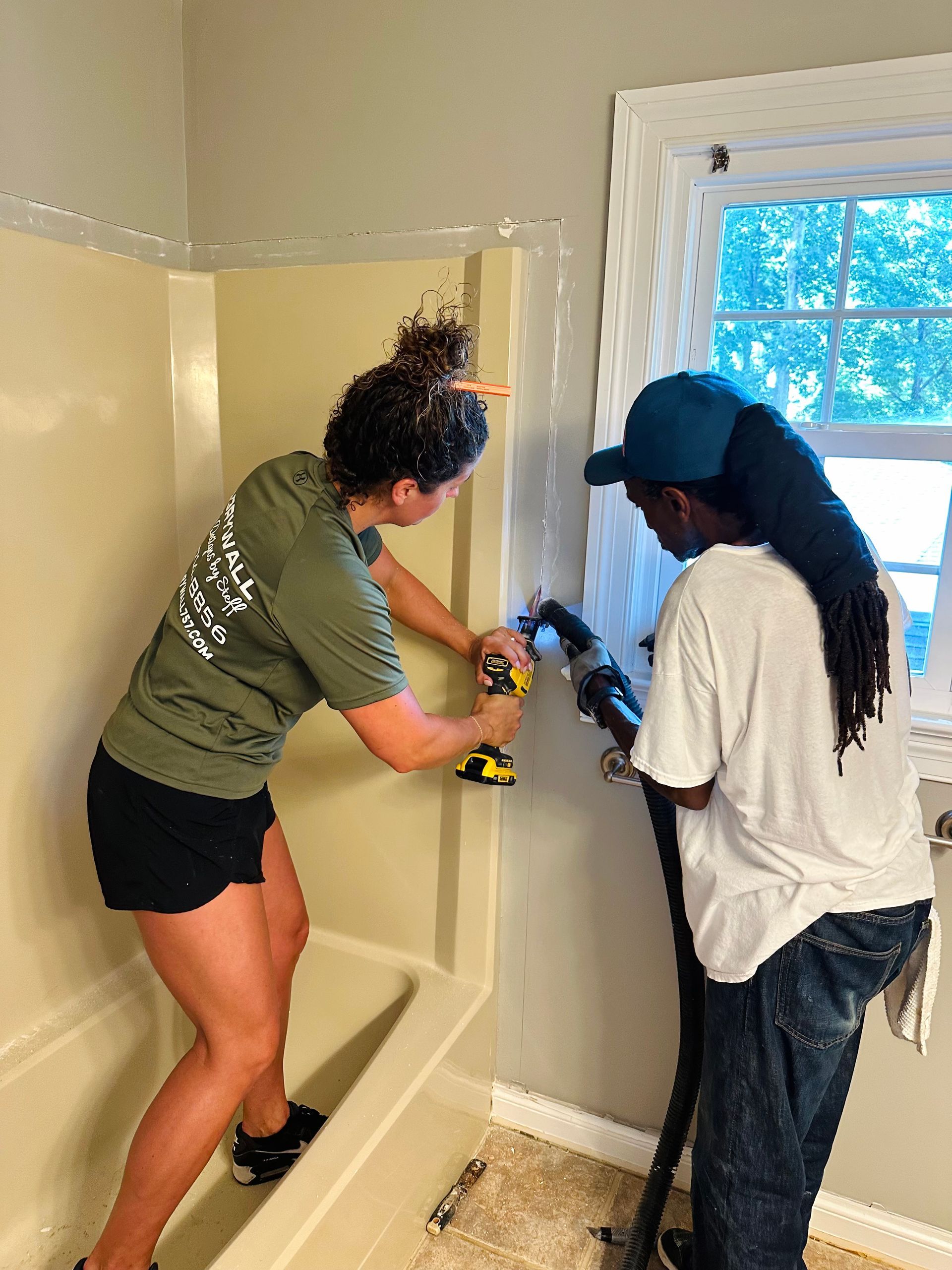Two people renovating a bathroom: one drilling, the other using a vacuum. Inside with a window.