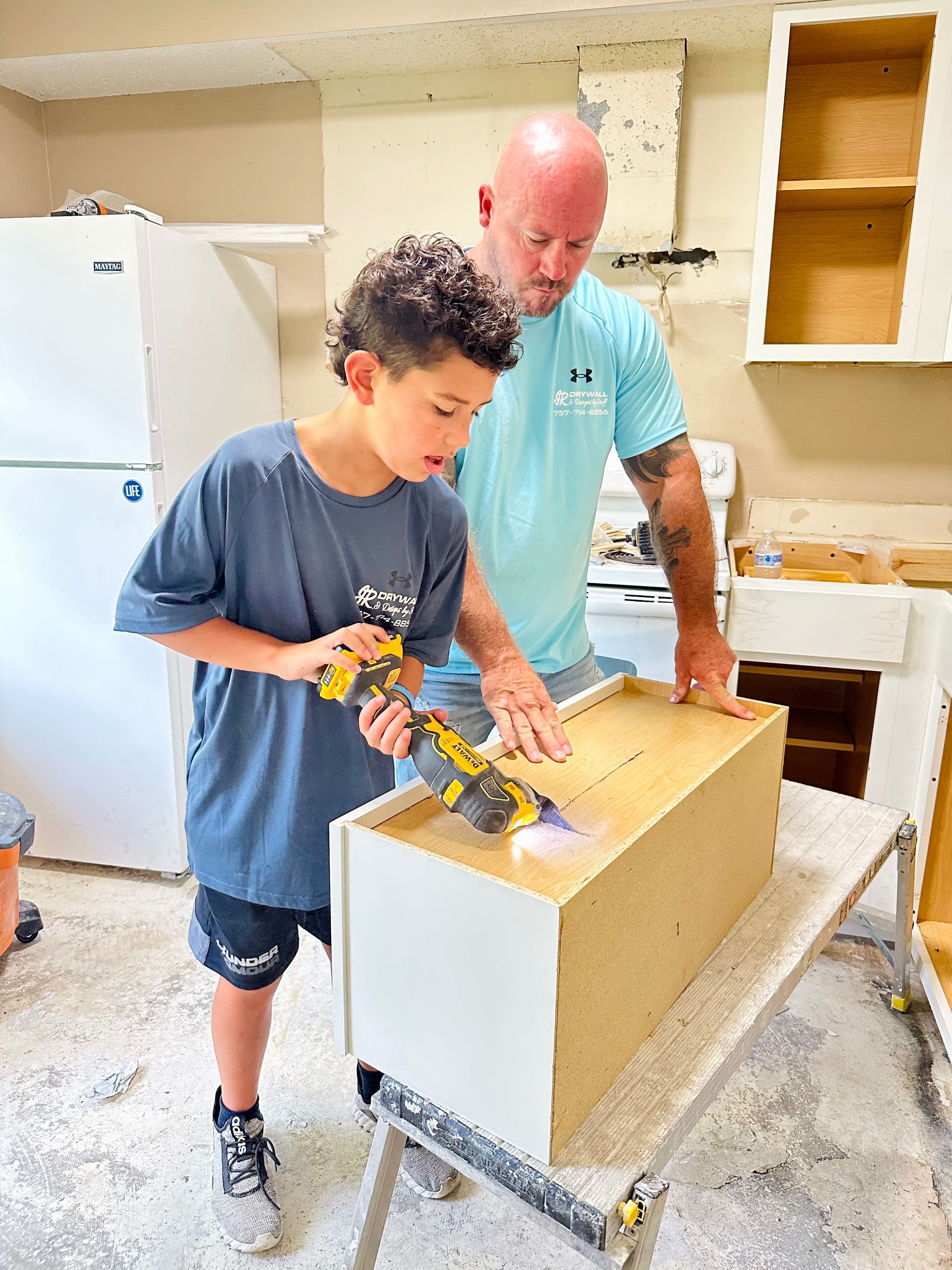 Man and boy sanding a cabinet in a kitchen. Boy uses a power tool while the man watches.