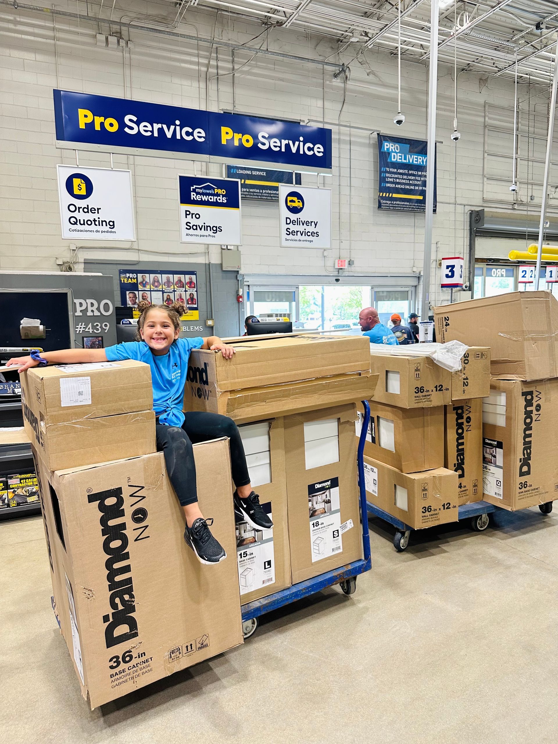 Girl sitting on boxes in a hardware store smiles with arms open, blue shirt, boxes say 