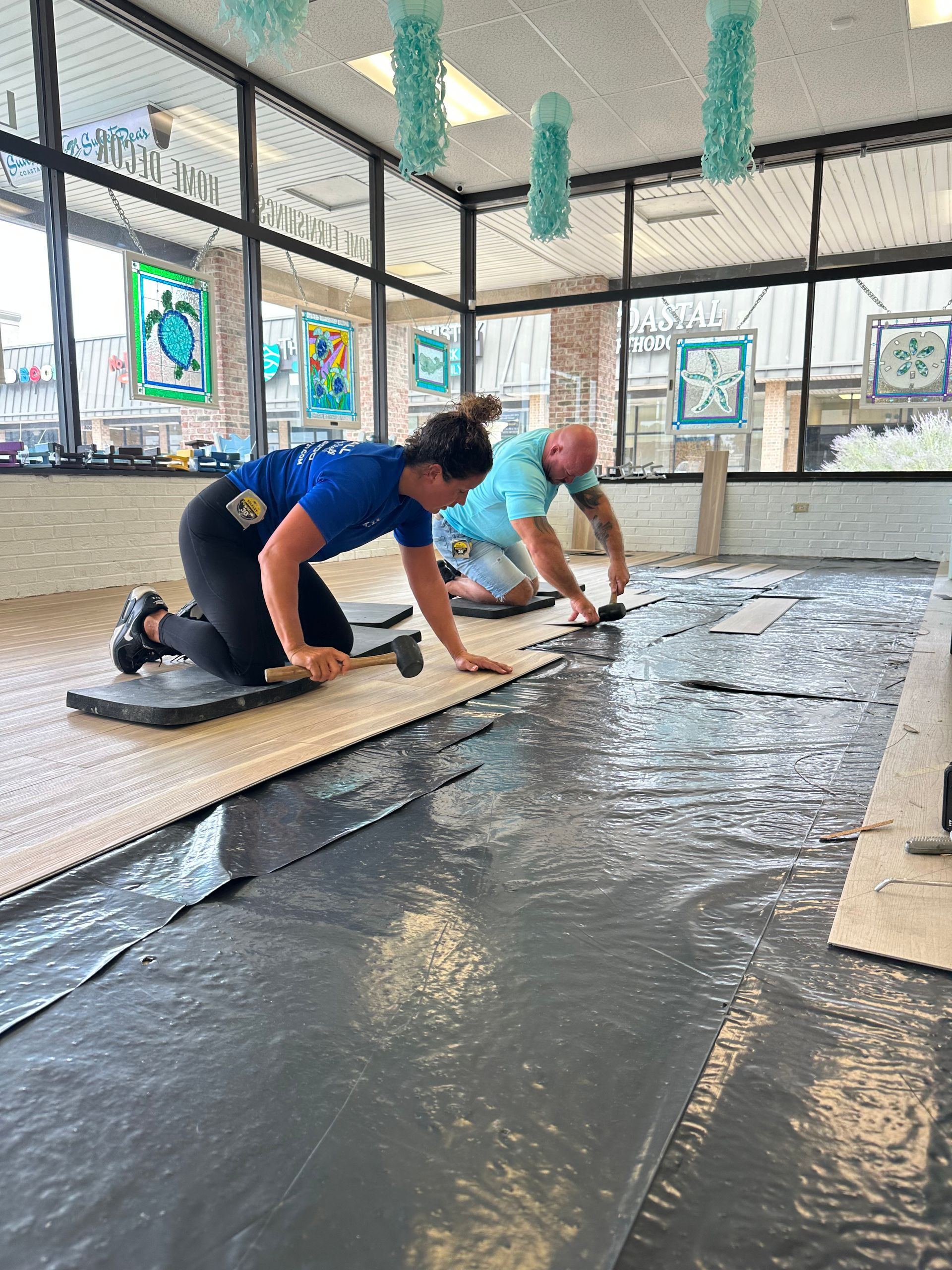 Two people installing flooring indoors; one kneeling, hammering, the other using a tool. Large windows in the background.