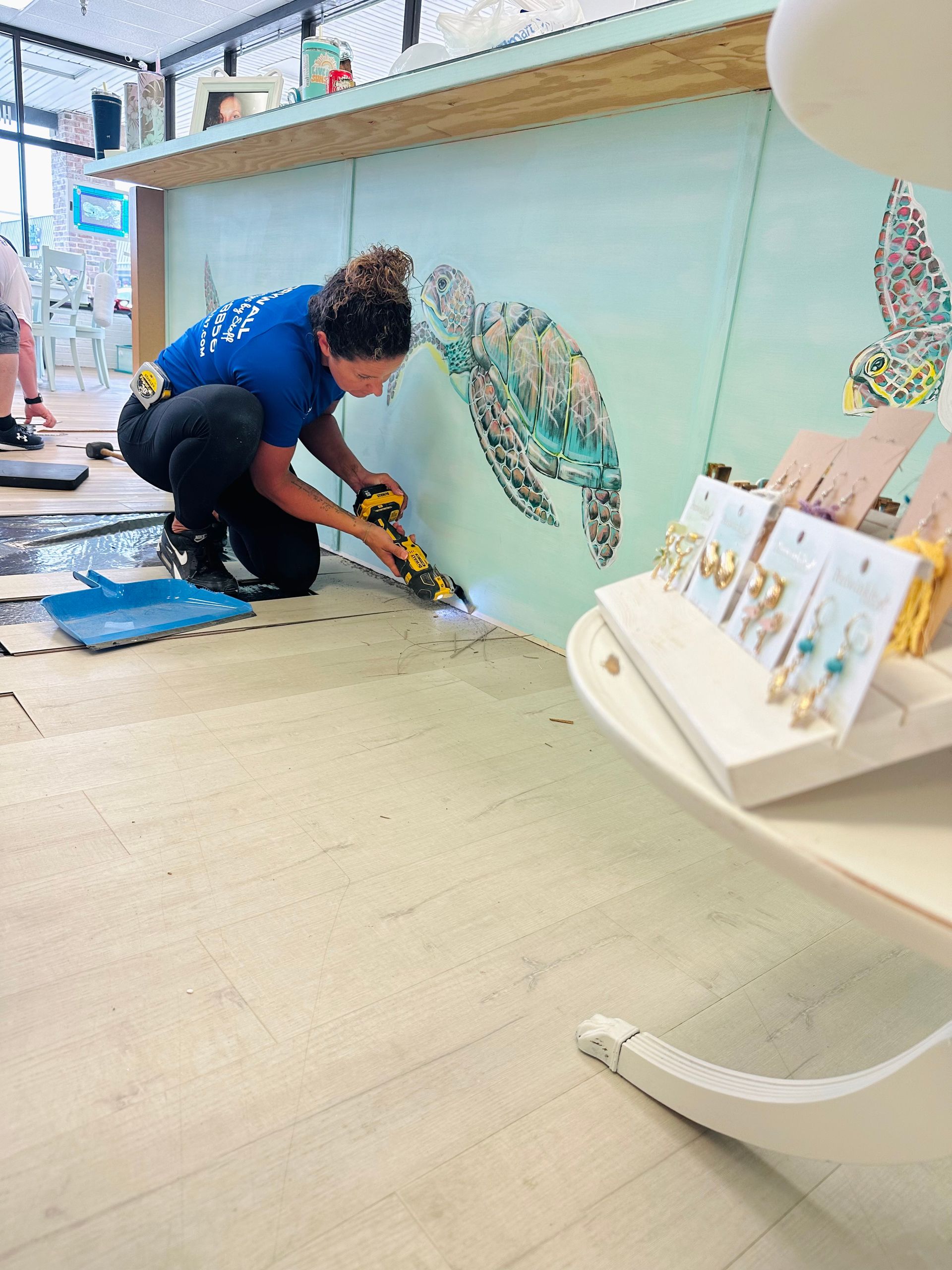 Woman in blue shirt uses a tool, working on wall mural of a turtle. Inside a store.