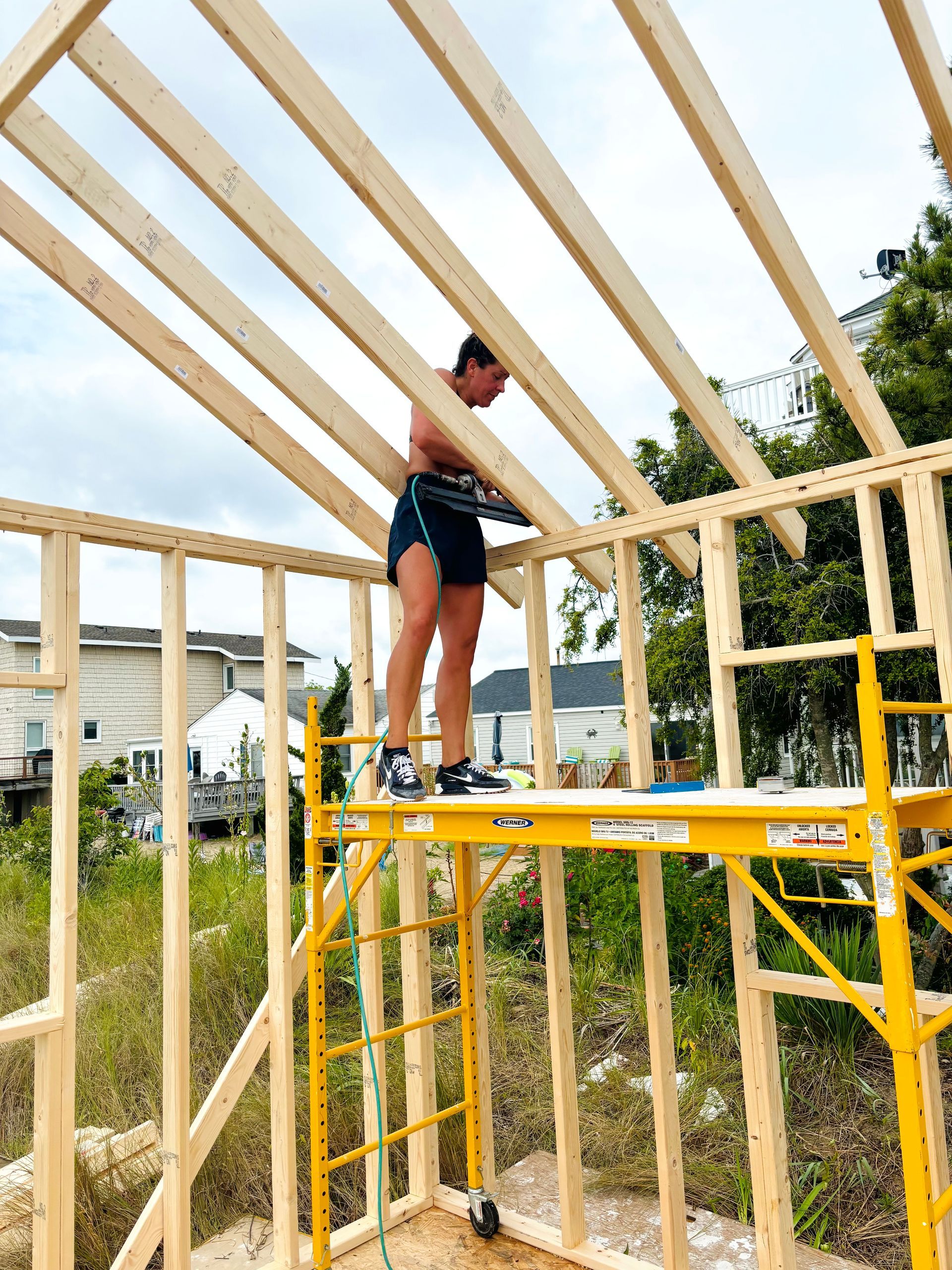 A person builds a wooden structure, standing on a yellow scaffold, outdoors.