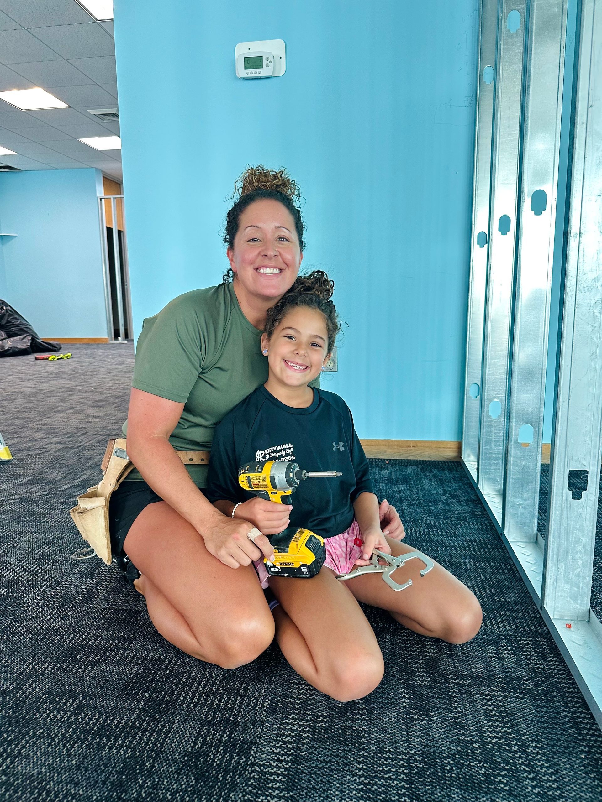 Woman and child kneeling by wall, holding a drill, smiling.