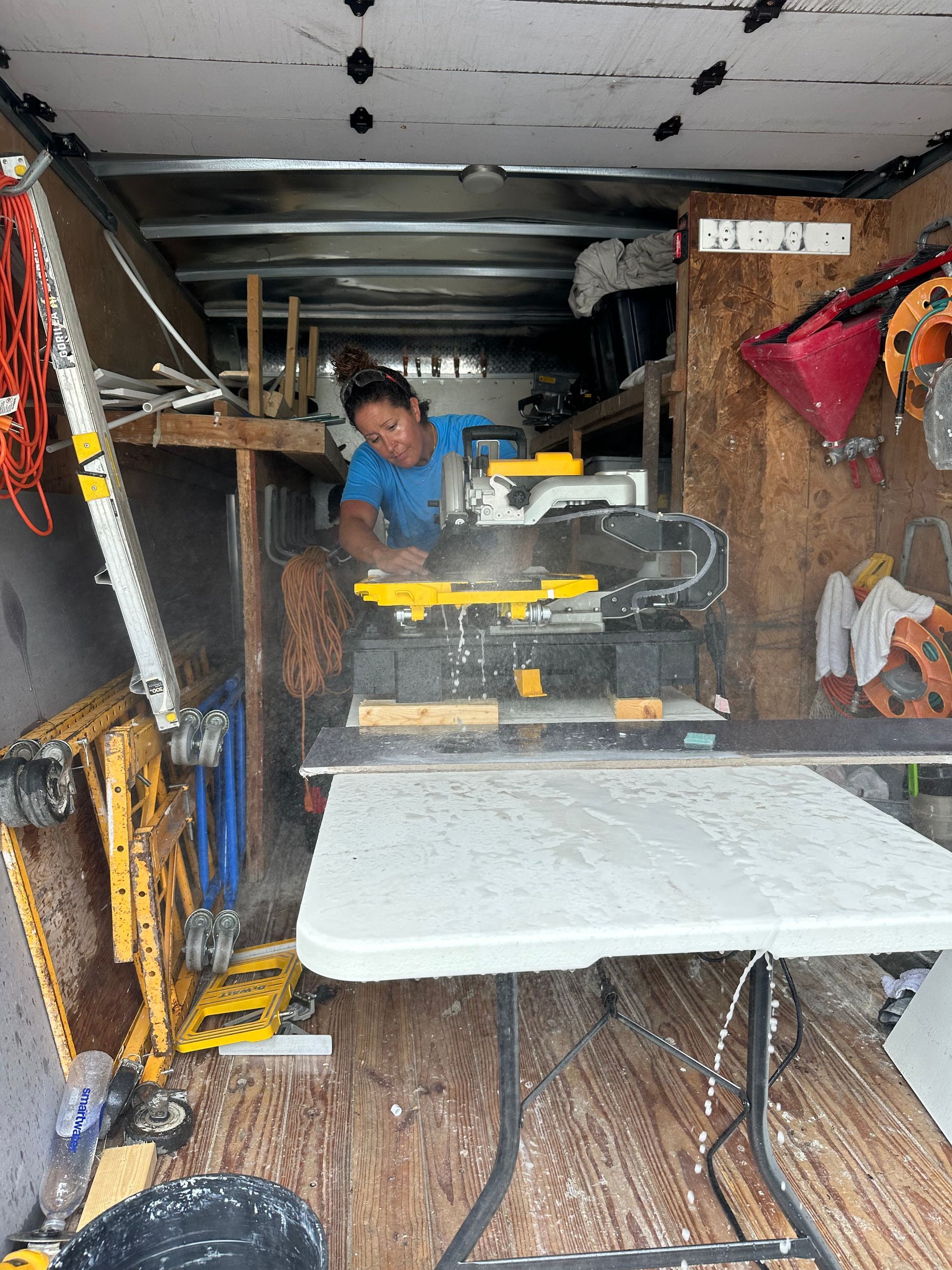 A person uses a yellow and black saw inside a trailer. Wood shavings and white table.