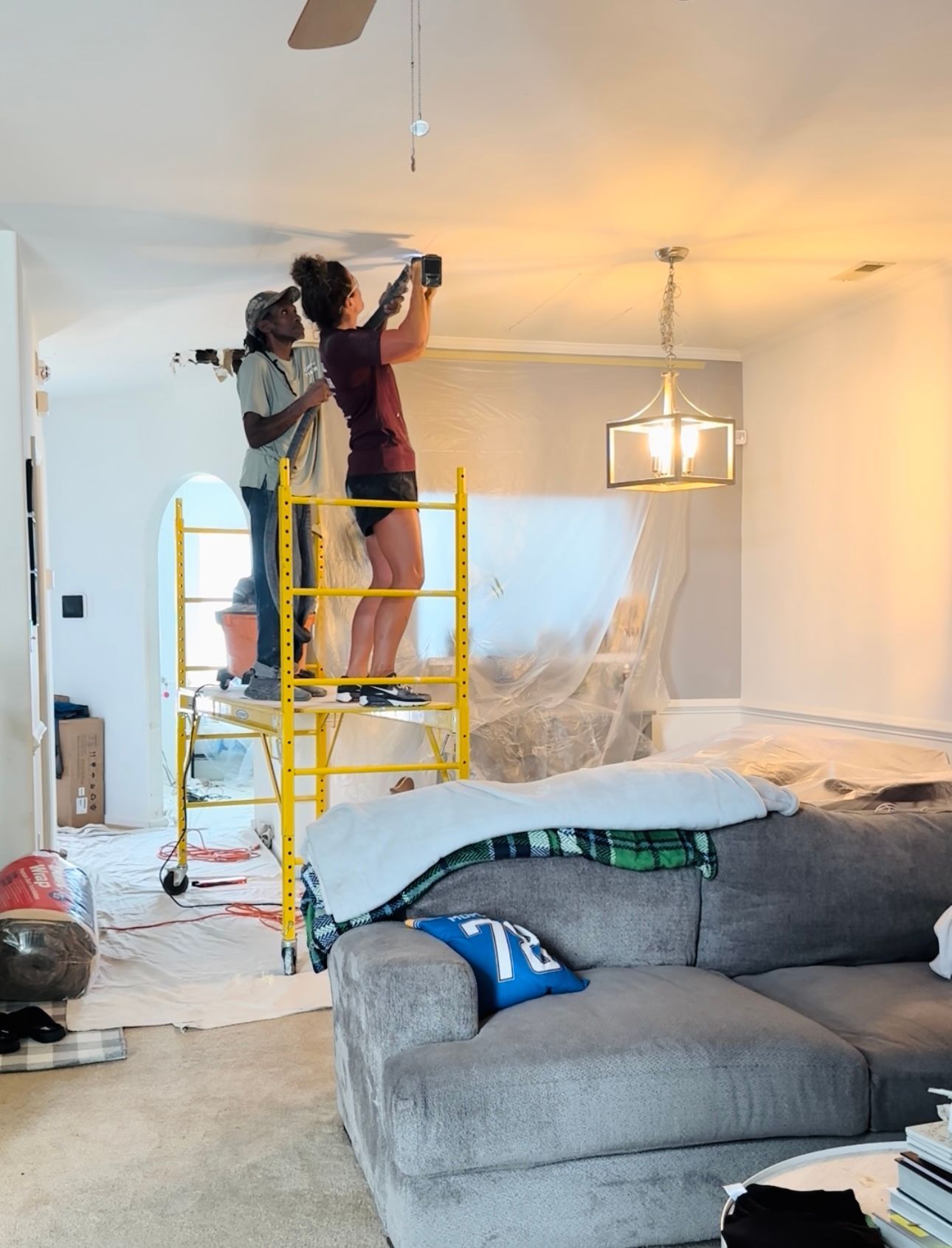 Two people painting a ceiling, standing on a yellow scaffold in a living room with furniture covered in plastic.