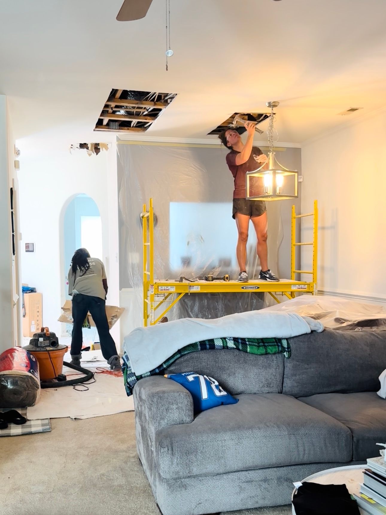 People renovating a room. One stands on scaffolding working on the ceiling, the other on the floor working too.