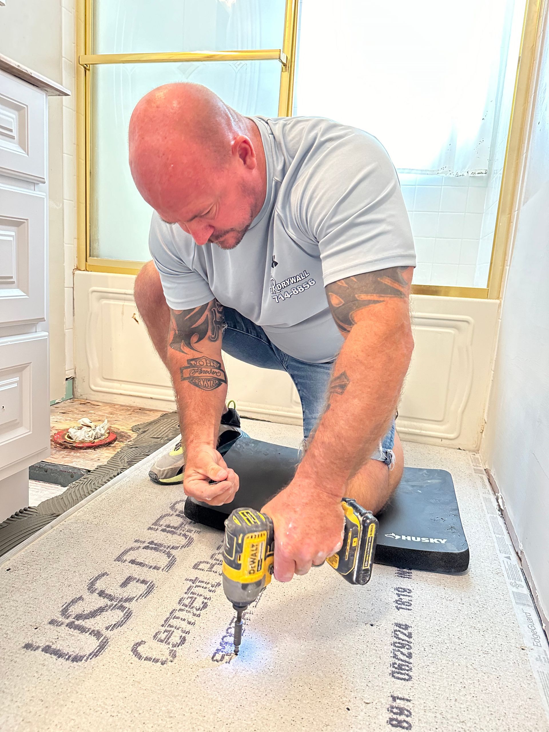 Man in bathroom, kneeling, using a drill on a white panel labeled 