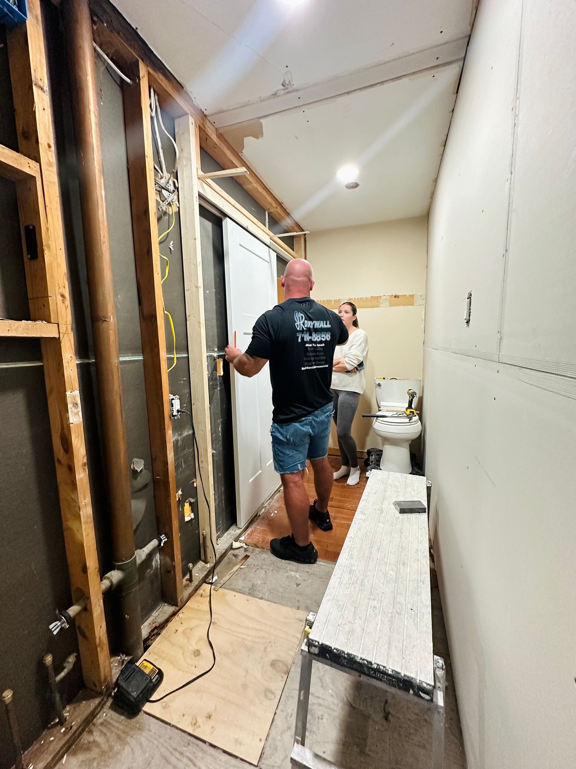 Man installing a door during bathroom renovation. White walls, wood framing, and a toilet visible.