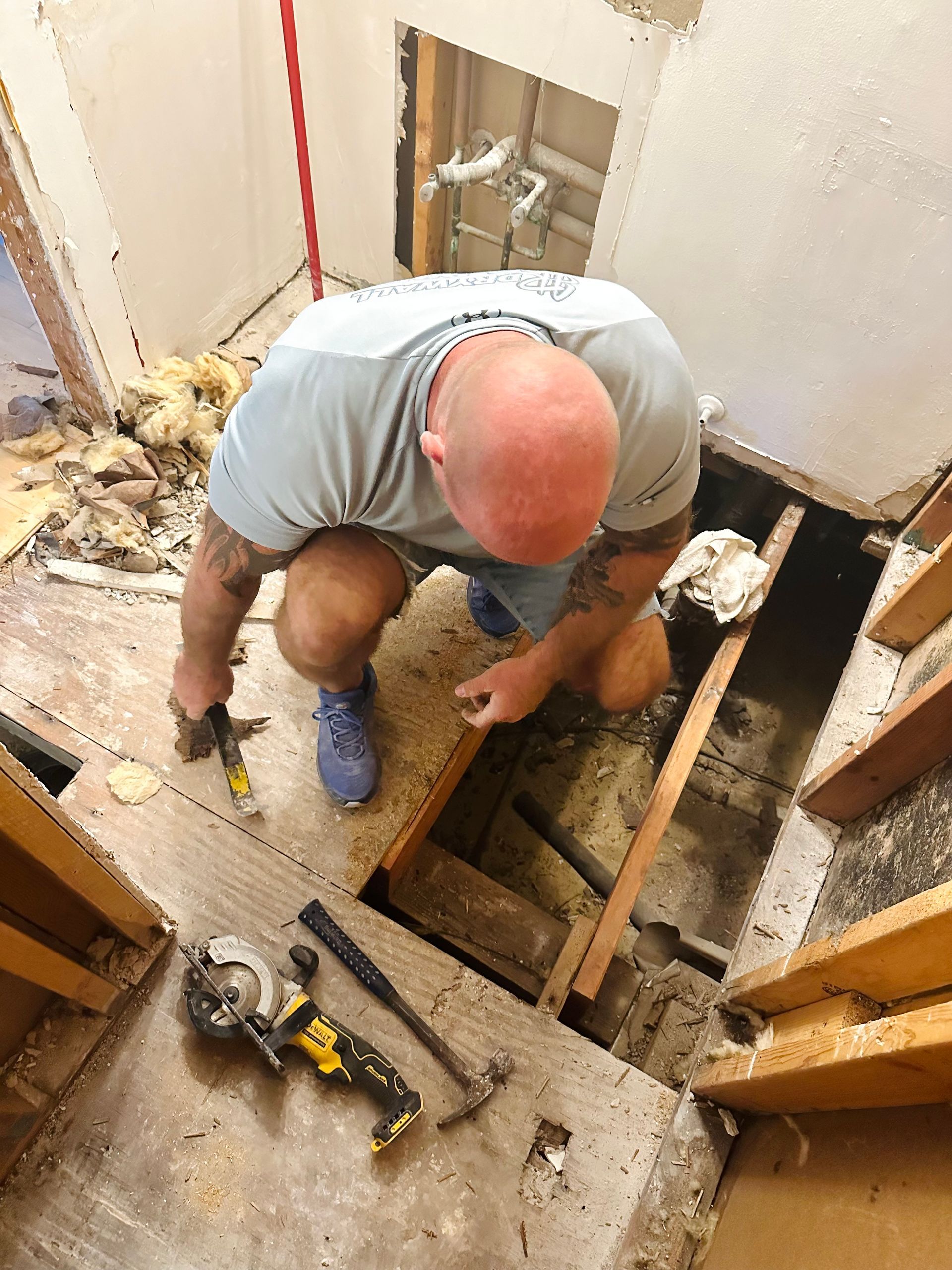 Man in grey shirt crouches on wooden floor, using tools for construction work.