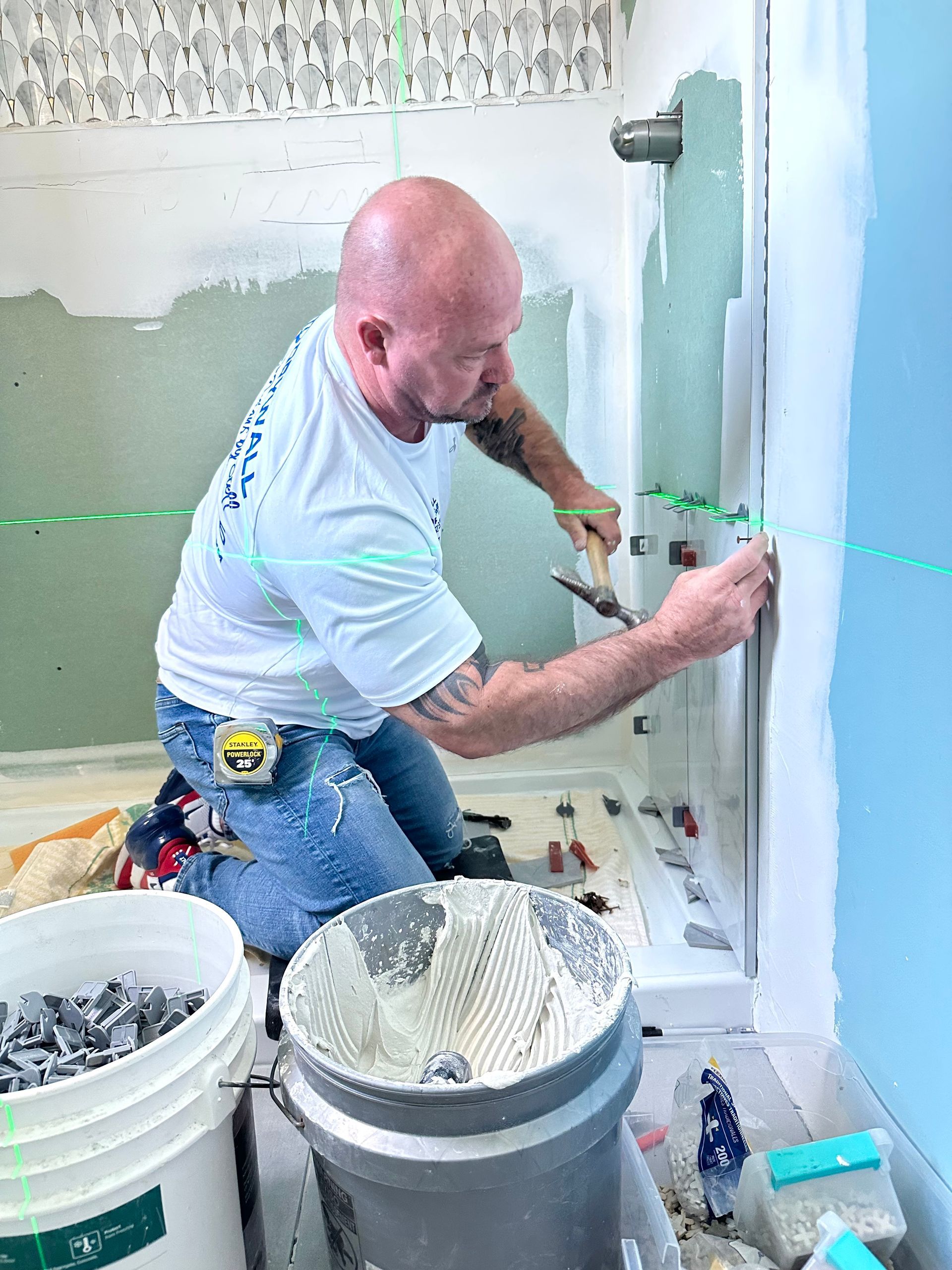 Man kneeling, tiling a shower. Green laser line guides tile placement. Tools and buckets visible.