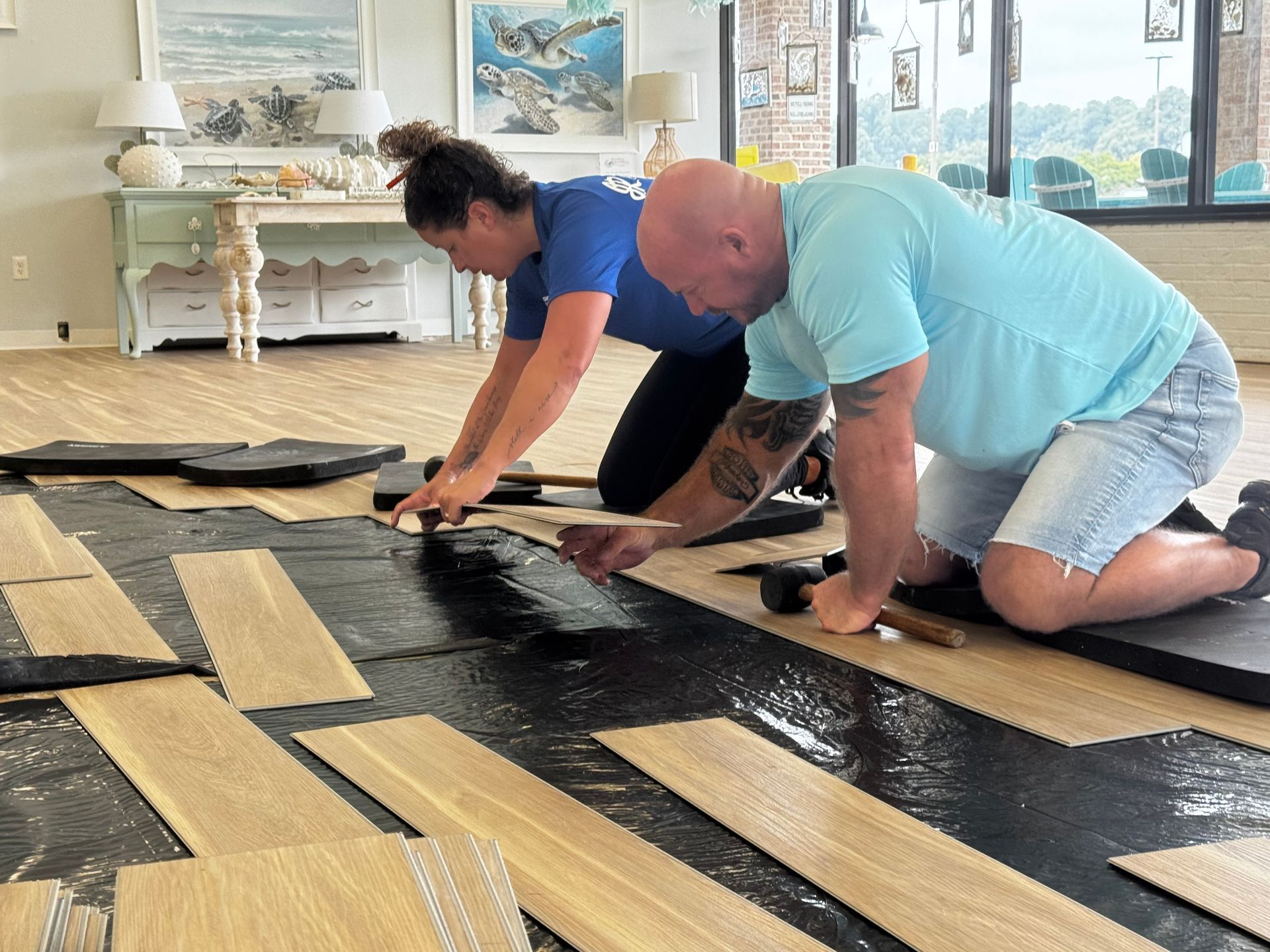 Two people kneeling, installing wood flooring in a room with coastal decor.