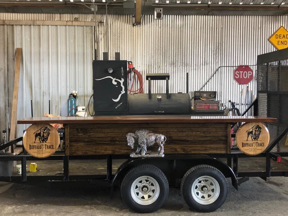 A mobile food trailer featuring a black barbecue smoker, a wooden counter, and a Buffalo Trace logo, parked in a garage.