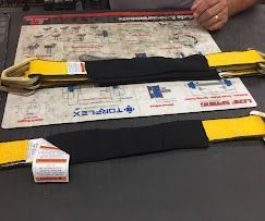 Two yellow nylon lifting slings with black protective wear pads and metal hardware, lying on a technical diagram mat.