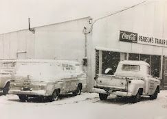A black and white photo shows two snow-covered vehicles parked in front of a building with a Coca-Cola sign.