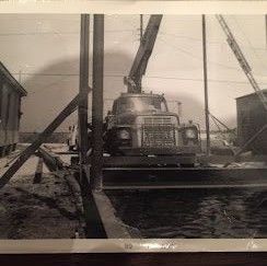 A vintage black and white photo of a large crane truck positioned between two buildings on a construction site.