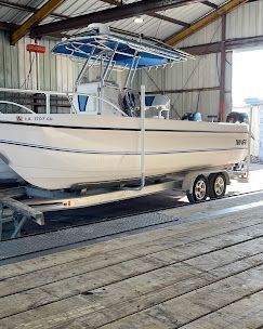 A white center-console boat with a blue T-top, mounted on a dual-axle trailer inside a covered boat storage facility.