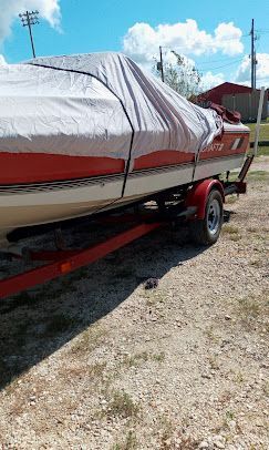 A red and white boat with a grey cover sitting on a trailer on a gravel lot under a partly cloudy sky.